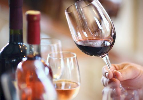 A hand holds a red wine glass beside a bottle and another glass, like a cozy tasting moment.