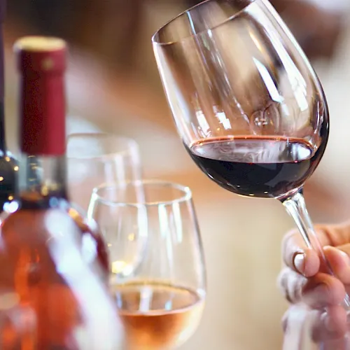 A hand holds a red wine glass beside a bottle and another glass, like a cozy tasting moment.