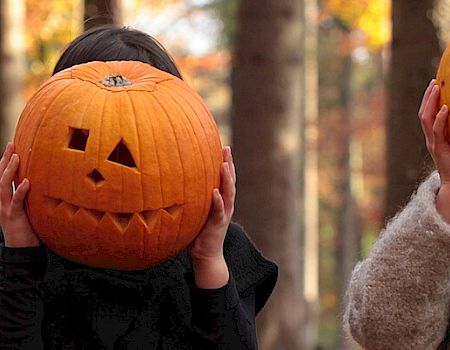 In a fall forest, three people in coats hold carved jack-o’-lantern pumpkins over their faces.