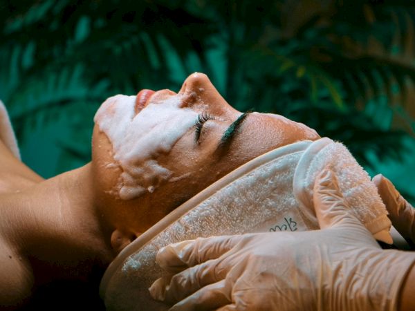 A facial treatment: a woman reclines while a therapist&rsquo;s gloved hands gently massage her face with cream.