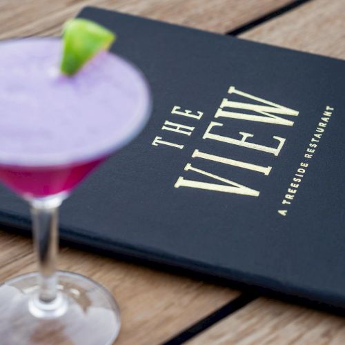 A purple cocktail with a lime wedge beside a dark menu with "The View - A Rooftop Restaurant" written on it, placed on a wooden surface.