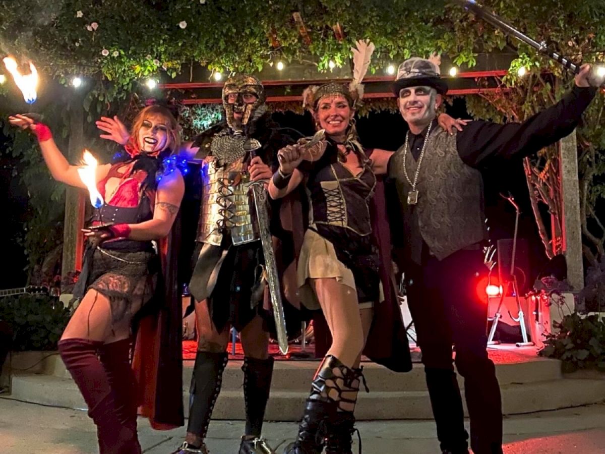 Four people in Halloween costumes pose outdoors at night, with one holding fire and the others in detailed, themed outfits.