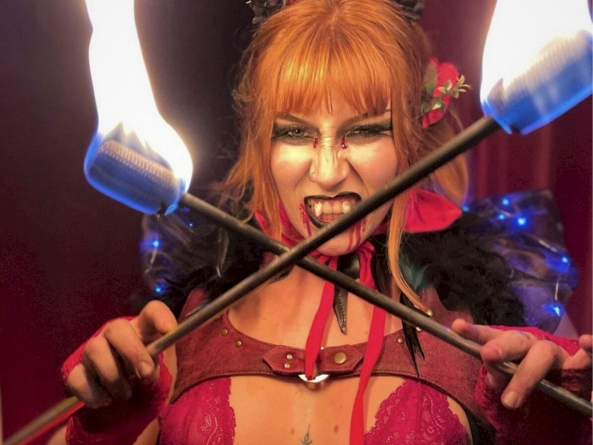 A person with intense makeup and costume holds crossed fire batons, creating a dramatic and fiery scene.