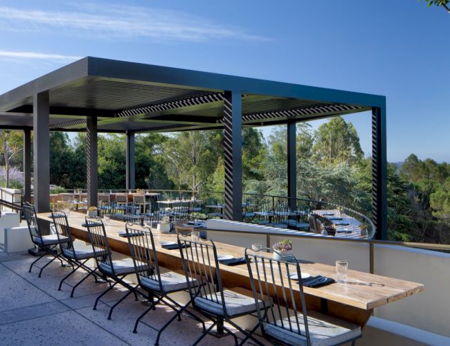 This image shows an open-air dining area with long wooden tables and chairs, partially covered by a pergola, with a view of trees and nature beyond.
