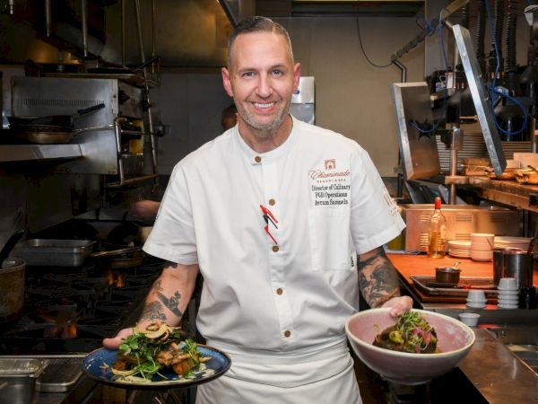 A smiling chef in a professional kitchen holds two plated dishes&mdash;one with greens and meat&mdash;posing by open stoves and cookware.