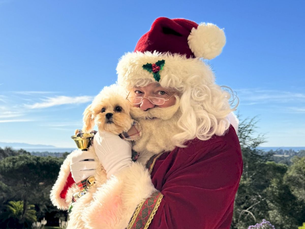 Santa in a red velvet suit holds a fluffy white dog outdoors under a blue sky, with a small golden cup in hand.