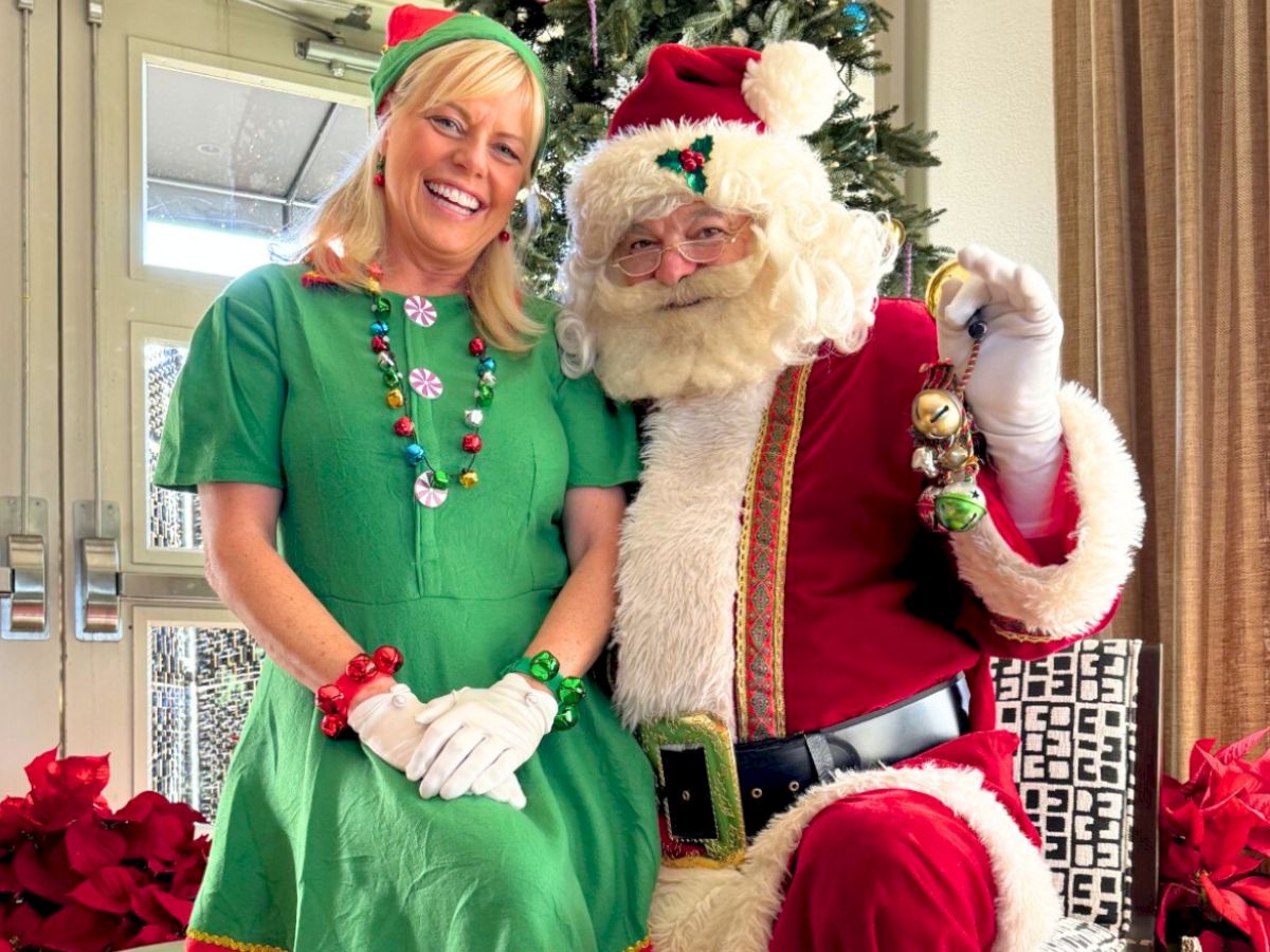 A smiling woman in a green Christmas elf outfit stands beside Santa Claus, posing in front of a decorated Christmas tree.