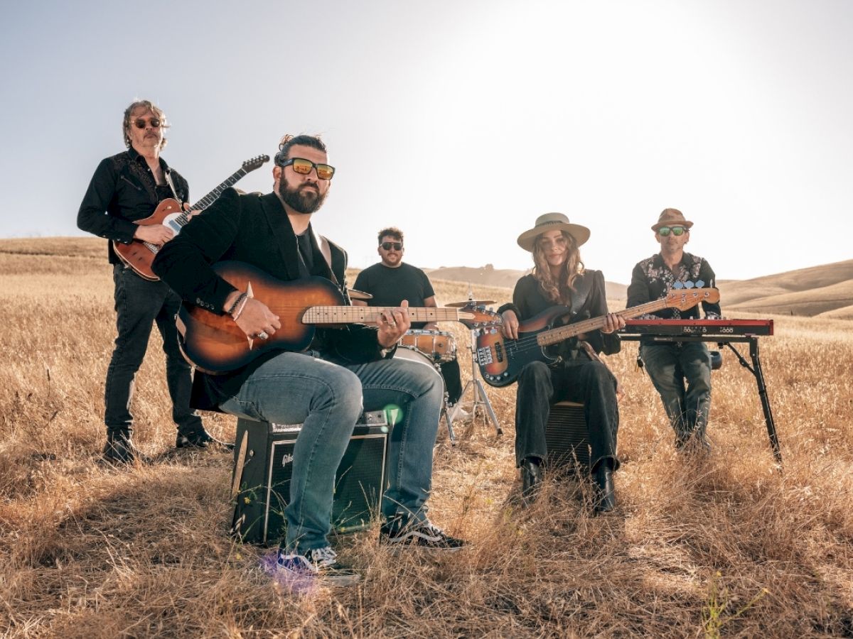 A country band with guitars and a keyboard performs in a sunny dry field of rolling hills.