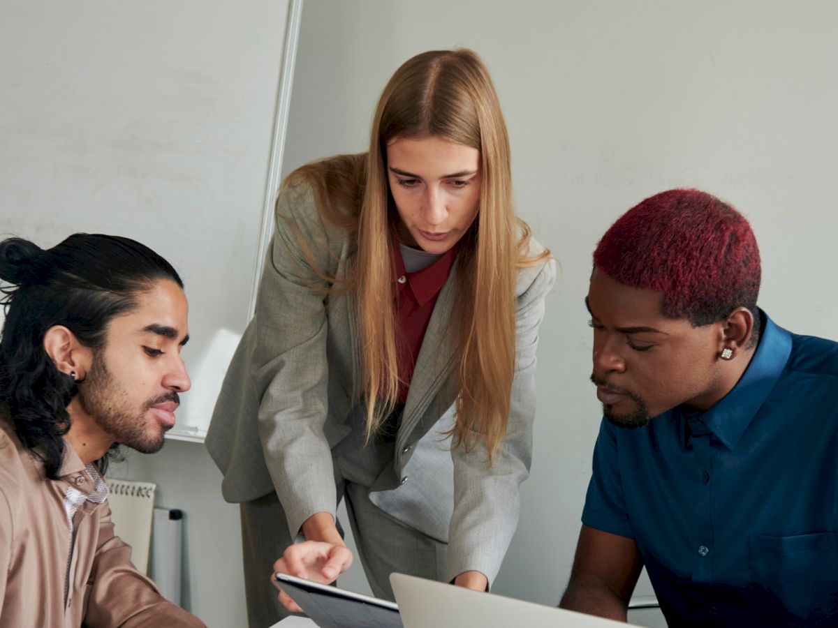 Three people are collaborating at a desk, with one pointing at a tablet and others looking at a laptop, in a meeting room.