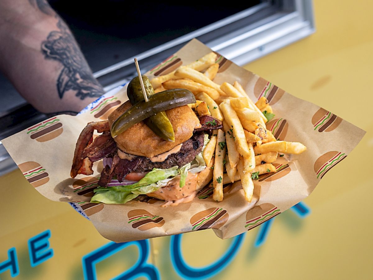 A tattooed arm holds a burger with pickles and fries on a paper tray in front of a yellow food truck.