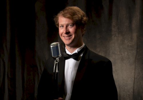 A person in formal attire stands in front of a vintage microphone against a dark backdrop, smiling for the camera.
