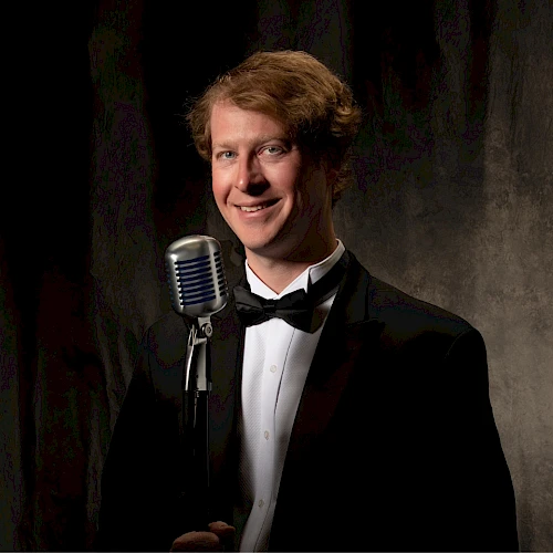 A person in formal attire stands in front of a vintage microphone against a dark backdrop, smiling for the camera.