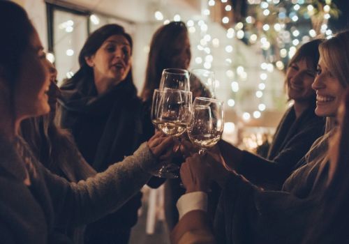 A group of people smiling and raising glasses in a toast, surrounded by festive lights in a cozy, dimly lit setting.