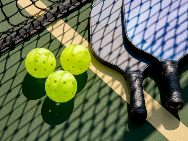 The image shows pickleball paddles, bright yellow pickleballs, and a net on a court.
