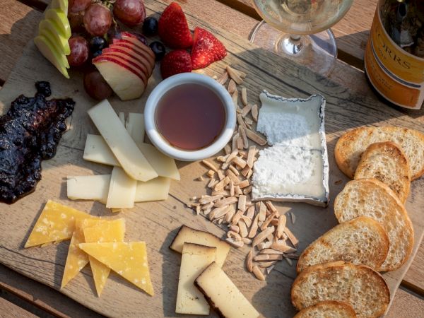 A rustic cheese board with a variety of cheeses, nuts, dried fruit, crackers, bread slices, a dark spread, and a small cup of tea or sauce on a wooden table.