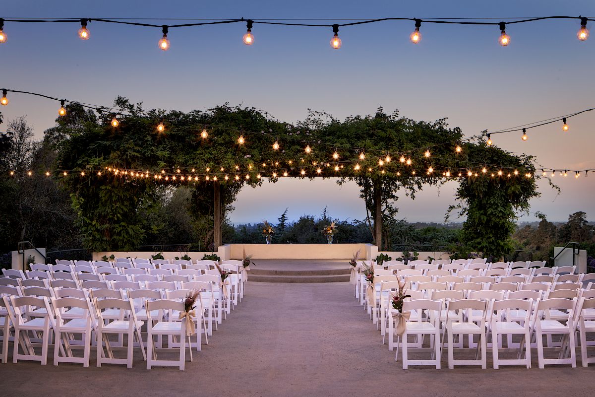 Outdoor wedding setup with white chairs and floral decorations under string lights and greenery, set against a scenic twilight backdrop.