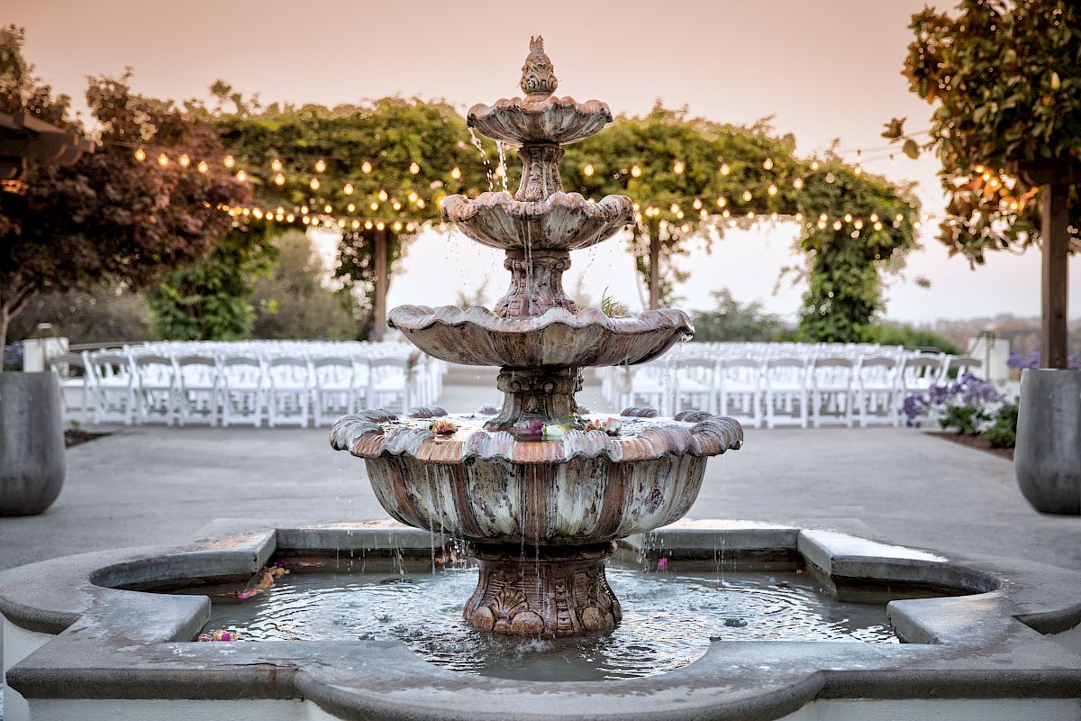 A tiered fountain is in the foreground with a pergola, string lights, and rows of white chairs in the background, suggesting an event.