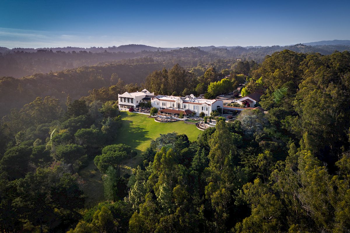 Aerial view of a large mansion surrounded by lush forests and hills, with a well-manicured lawn in the foreground.