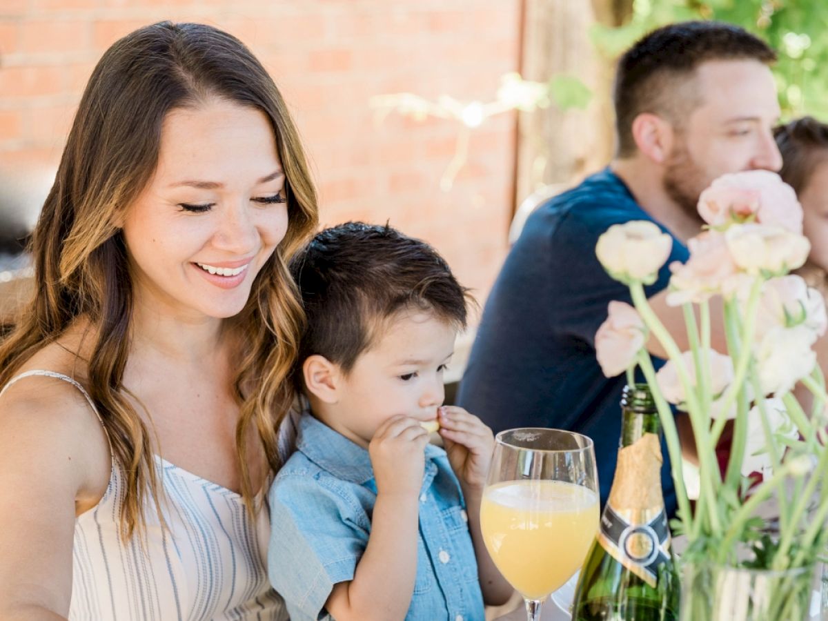 A family enjoys an outdoor meal. A woman and child are smiling together, a drink and flowers are on the table.