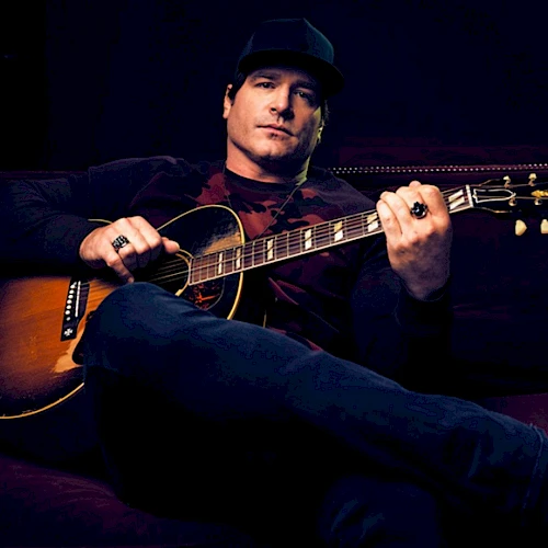 A person lounging on a sofa, holding an acoustic guitar, wearing casual clothing, and a cap, set against a dark background.