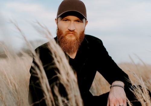A person with a beard and cap is sitting in a field of tall grasses, looking directly at the camera under a partly cloudy sky.