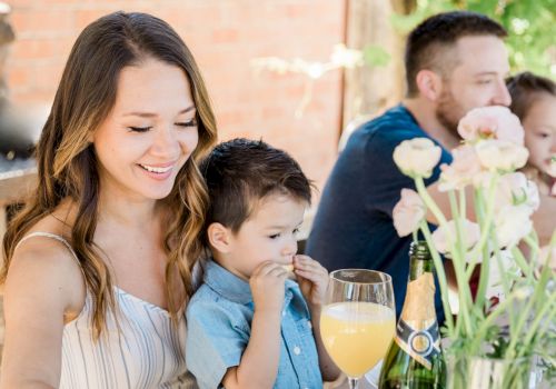 A woman and child are smiling at a table with flowers and drinks, joined by another adult and child in the background.