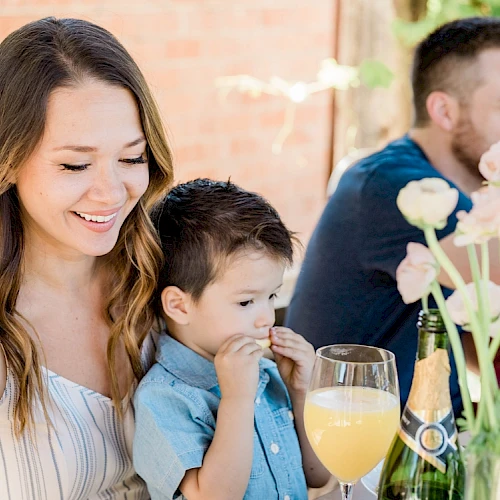 A woman and child are smiling at a table with flowers and drinks, joined by another adult and child in the background.