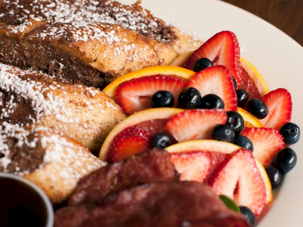 Slices of bread dusted with powdered sugar, garnished with strawberries, oranges, blueberries, and a piece of bacon on a plate.