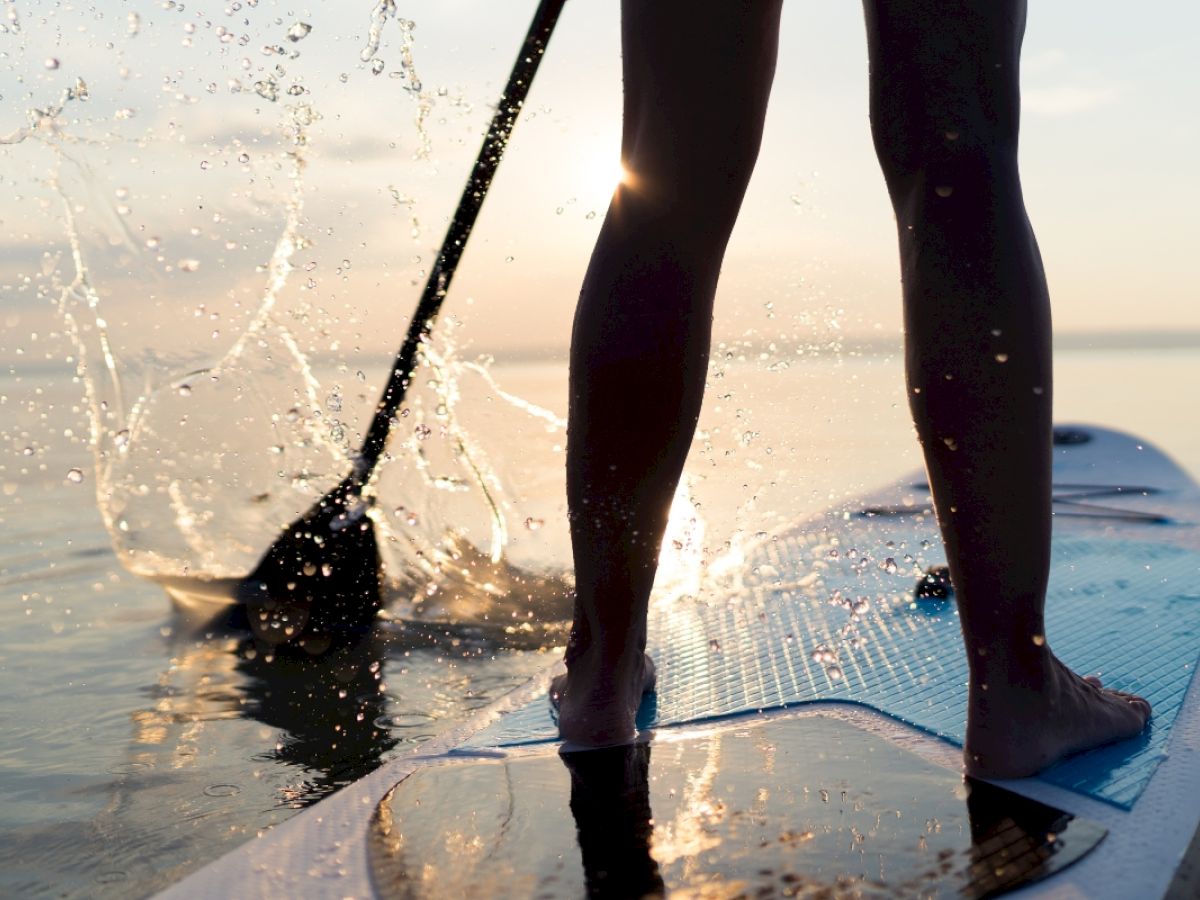 A person stands on a paddleboard, splashing water with a paddle, during a serene sunset on a calm body of water.