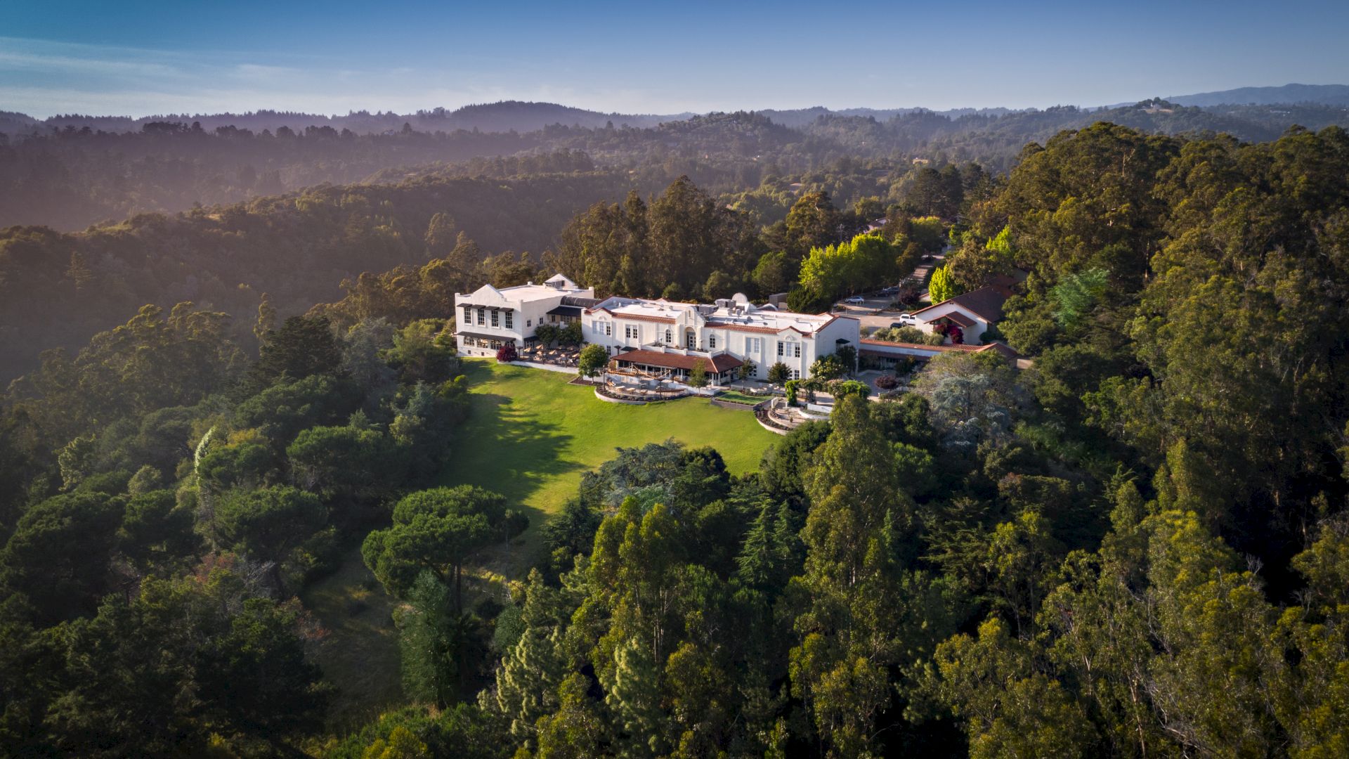 A large estate surrounded by lush greenery and rolling hills under a clear blue sky.