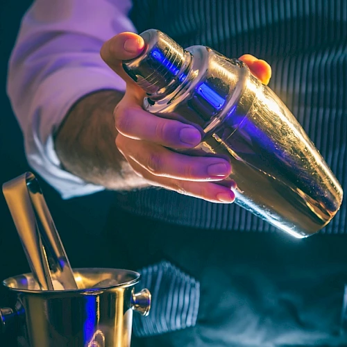 A person holding a shiny cocktail shaker above an ice bucket, possibly preparing a drink.