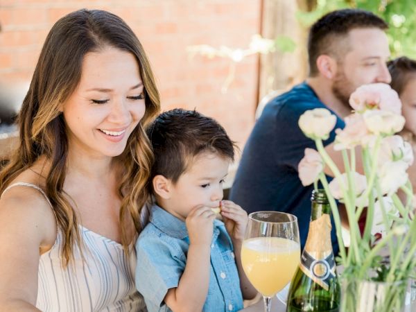 A woman and child are seated at a table with flowers, sharing a joyful moment, with drinks and another adult visible in the background.