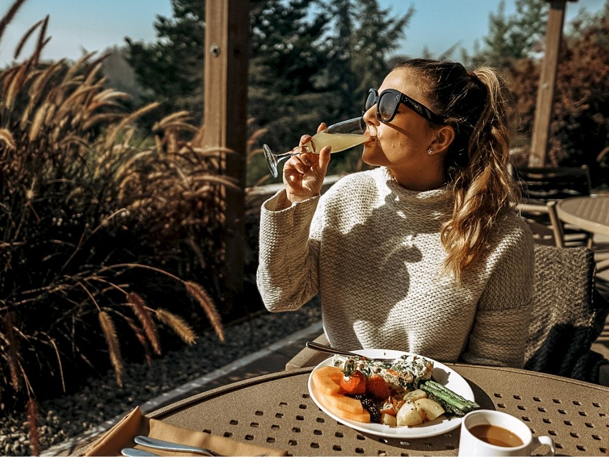 A person in sunglasses is enjoying a drink, with a plate of food and a cup of coffee on an outdoor table amid foliage.