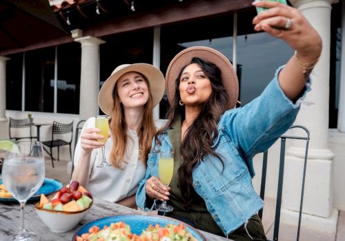 Two friends take a selfie while enjoying drinks and food on an outdoor patio, smiling and posing for the photo.