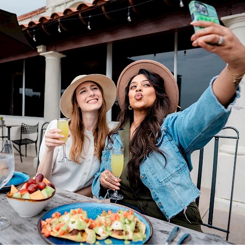 Two friends take a selfie while enjoying drinks and food on an outdoor patio, smiling and posing for the photo.