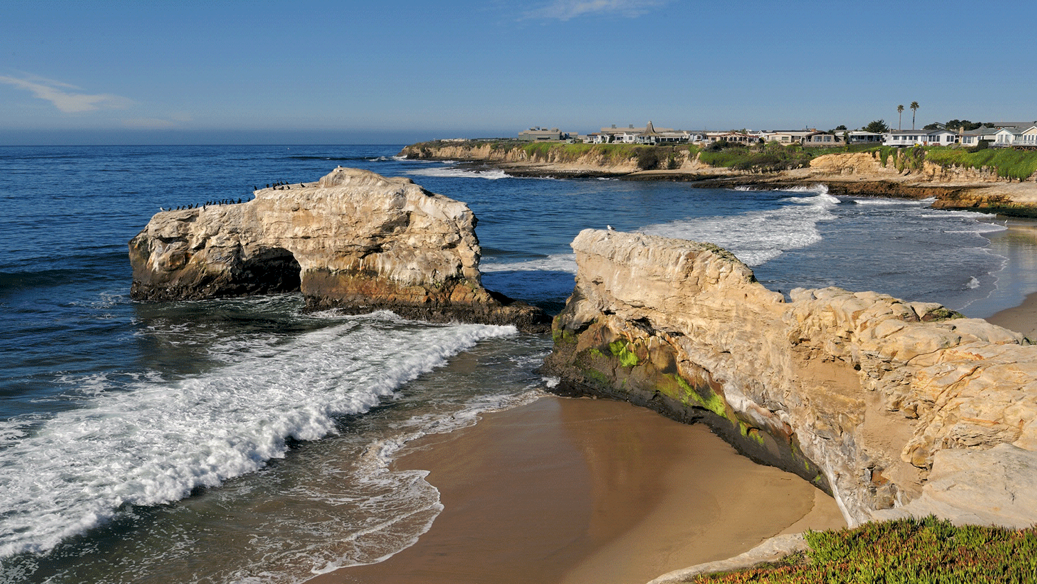 A coastal scene with rocky cliffs, a small sandy beach, ocean waves, and houses in the distance under a clear blue sky.