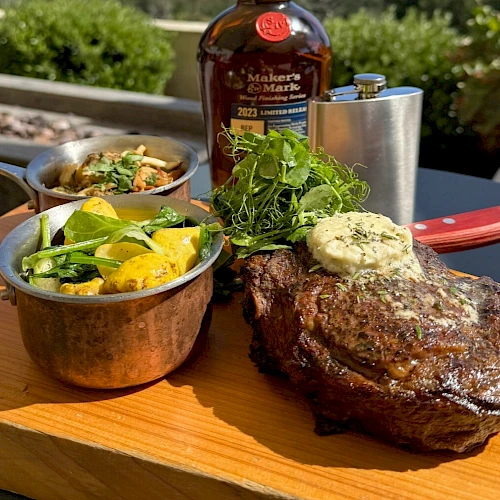 A grilled steak with herb butter, accompanied by vegetables in pots, and a bottle in the background, served on a wooden board.