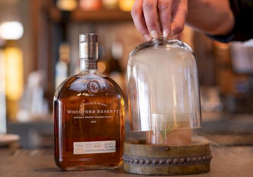 A bottle of bourbon and a cocktail under a glass dome with smoke, beside a hand lifting the dome in a bar setting.