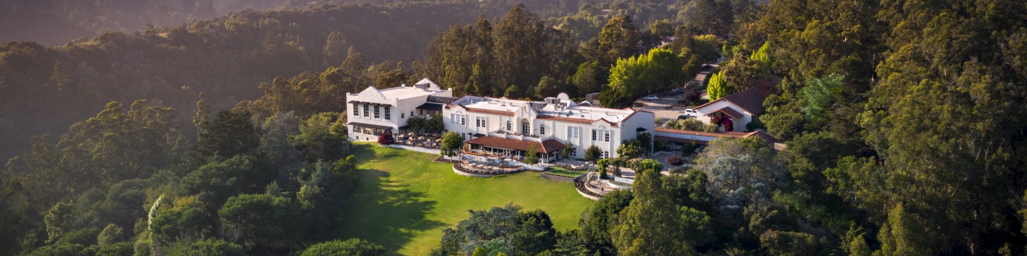 Aerial view of a white villa with red-tile roofs surrounded by lush greenery and rolling hills at golden-hour light.