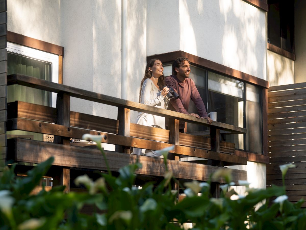 A smiling couple relaxes on a wooden balcony, sipping coffee and chatting in warm sunlight.
