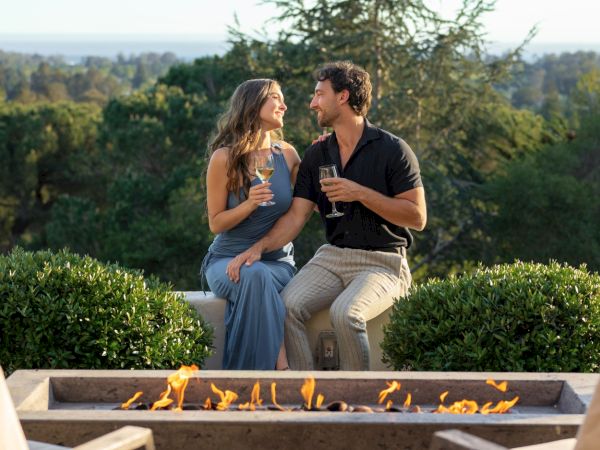 A couple sits by a fire pit on a patio, sharing drinks and smiling at each other against a scenic, tree-filled backdrop.