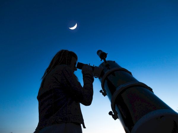A person is using a telescope to observe the sky, with a crescent moon visible in the background against a twilight sky.