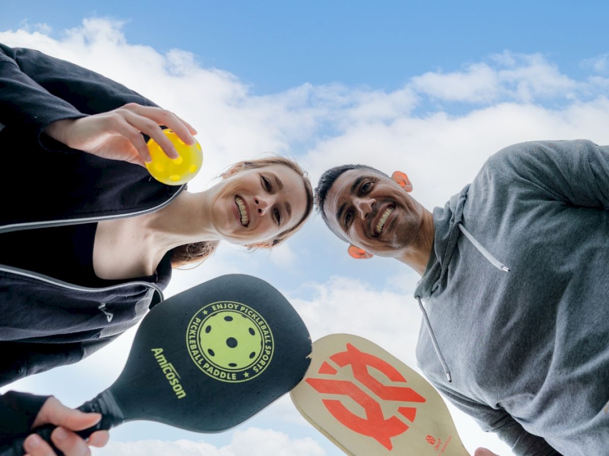 Two people are looking down, holding pickleball paddles and a ball, with a blue sky and clouds in the background.