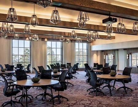 A bright conference room with round tables, black chairs, hanging pendant lights, carpet, and large windows—ready for meetings and events.