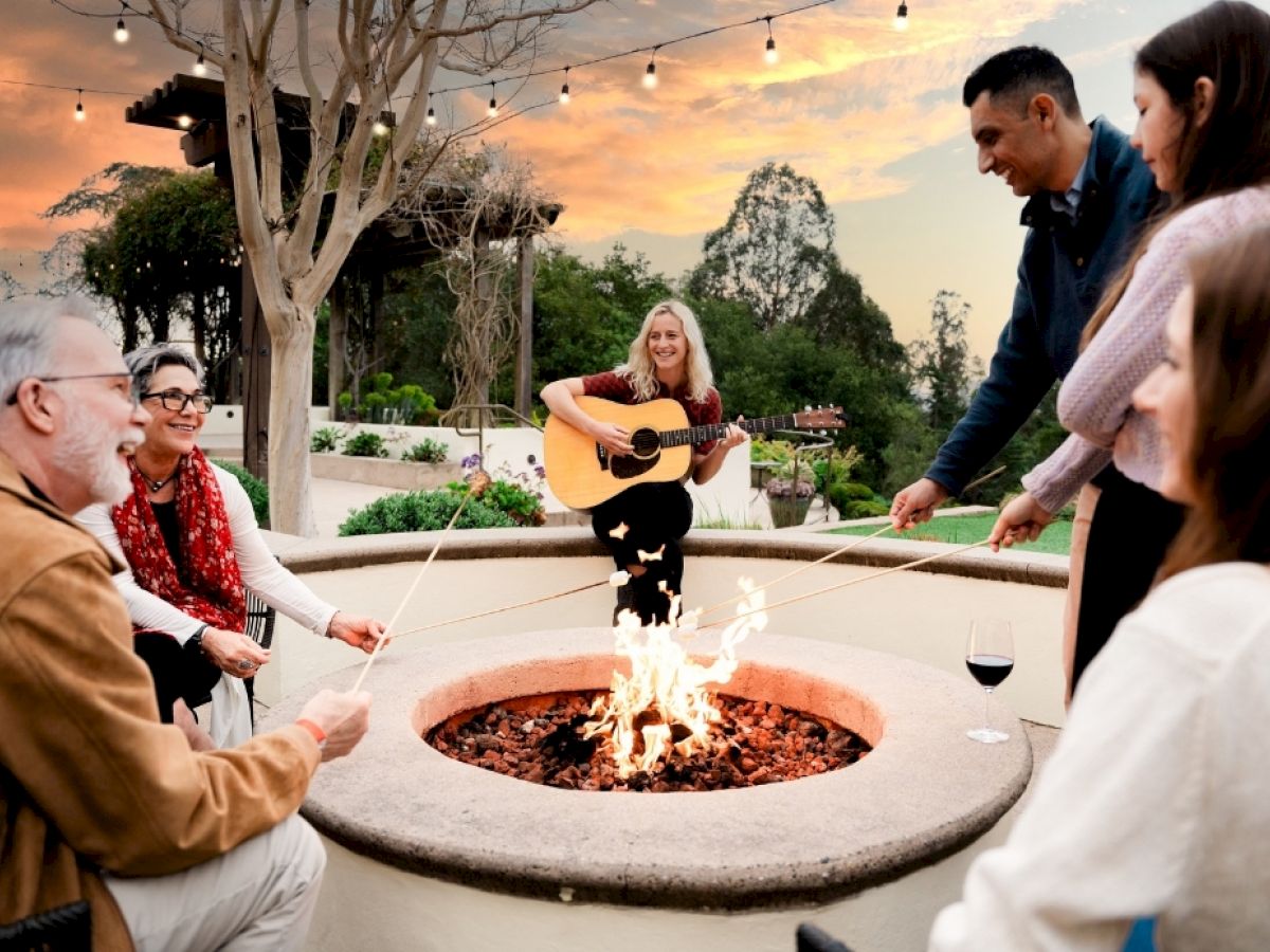 Friends gather around an outdoor fire pit as someone plays guitar at sunset, roasting with drinks in a cozy yard.