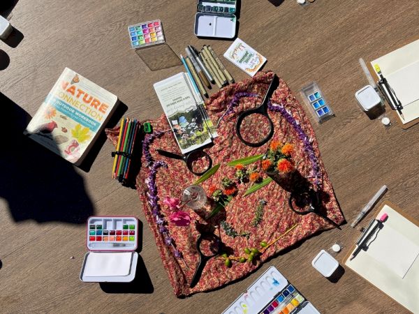 An overhead view of a painter&rsquo;s desk with an artwork print, colored pencils, watercolors, markers, brushes, and sketch materials.