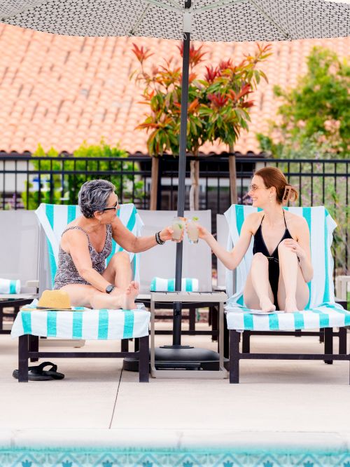 Two people relaxing by a pool give each other a high five on lounge chairs under an umbrella.