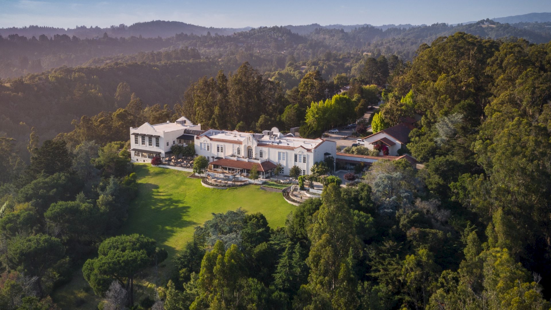 An aerial view of a large, luxurious mansion surrounded by lush greenery and hills with a well-maintained garden and trees.