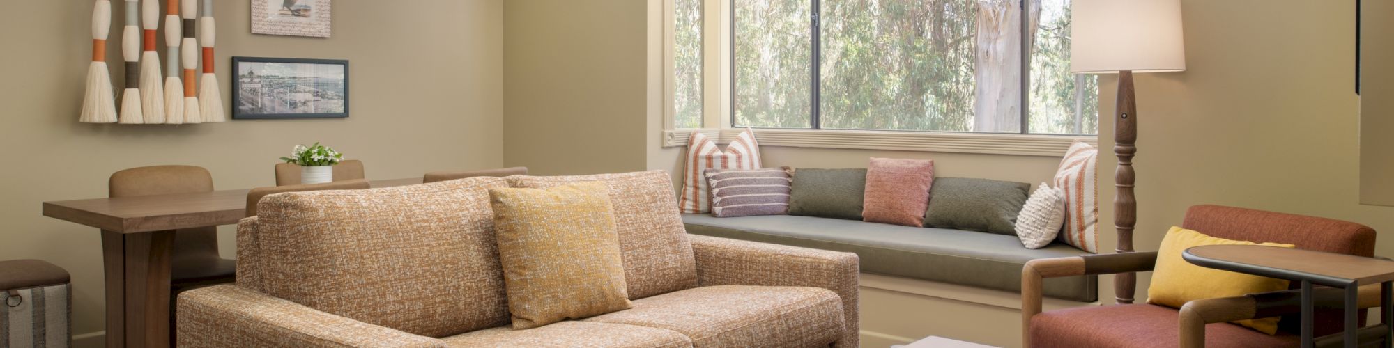 Bright modern living room with sofa, coffee table, armchairs, wall decor, and a large window with greenery outside.
