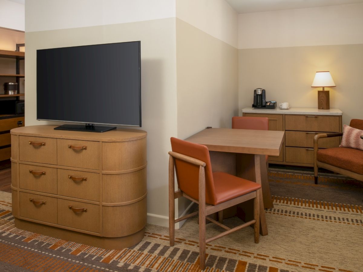 A cozy hotel living area with a wooden dresser supporting a large flat-screen TV, a wooden dining table and chairs, a kitchenette with a coffee maker, lamp, and sofa in warm, neutral tones.